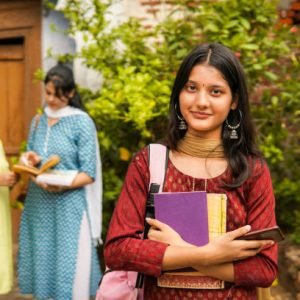 Portrait a beautiful indian female college student in salwar kameez and dupatta with backpack and a book in hand looking at the camera with confident smile and other female college students standing in the background. Rural Indian and education concept.